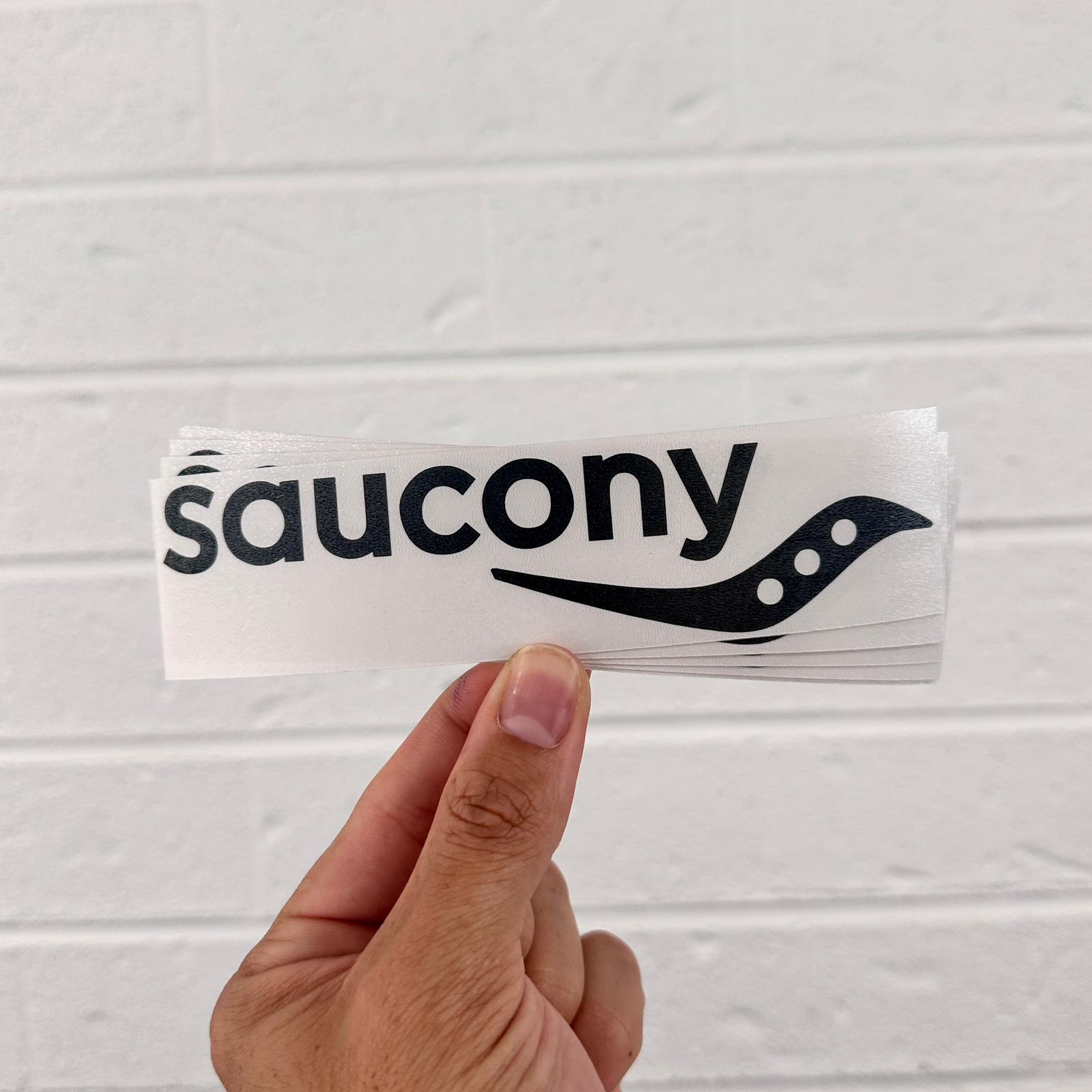 Hand holding a Saucony vinyl sticker against a light gray background