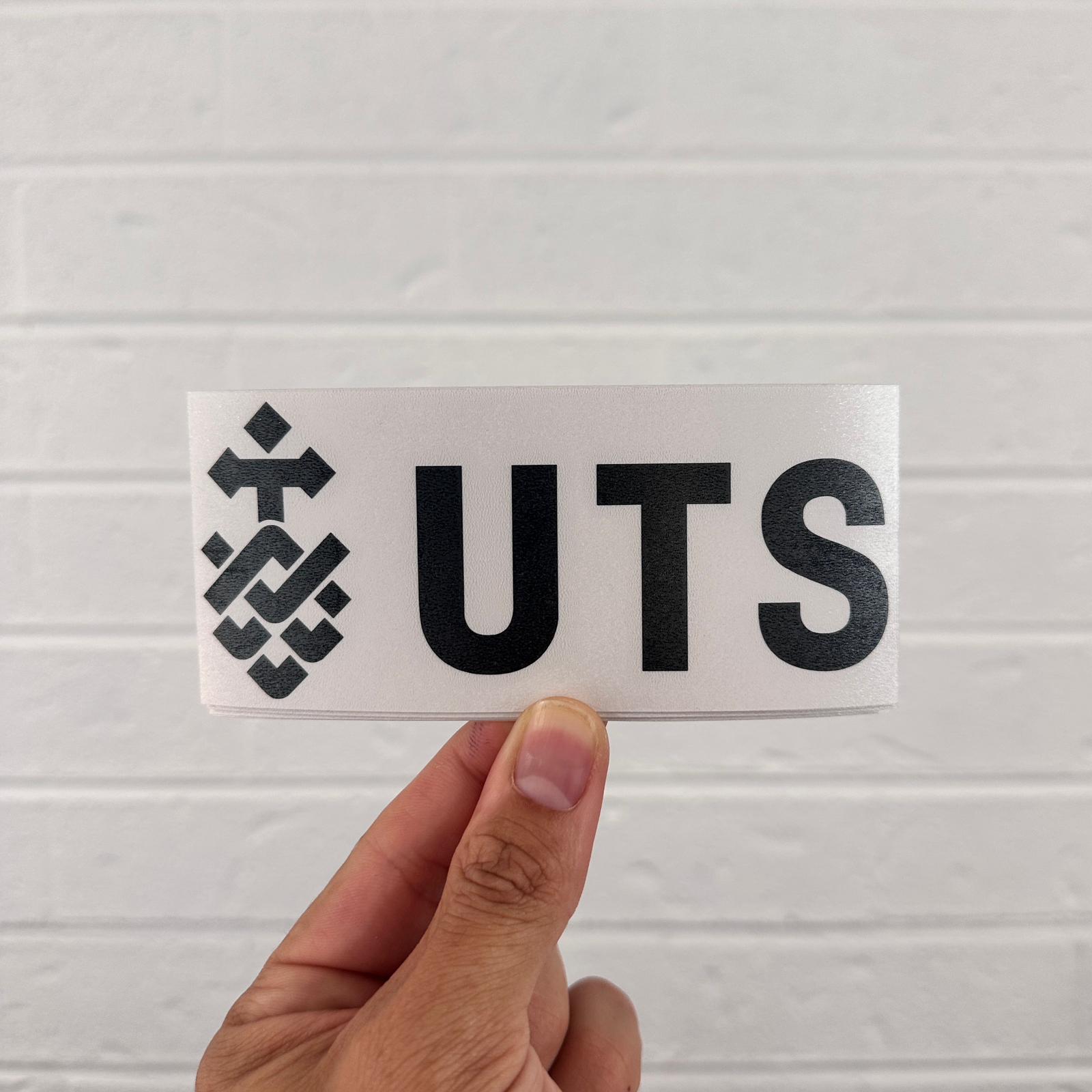 Hand holding a sticker with 'UTS' and a logo against a light gray background