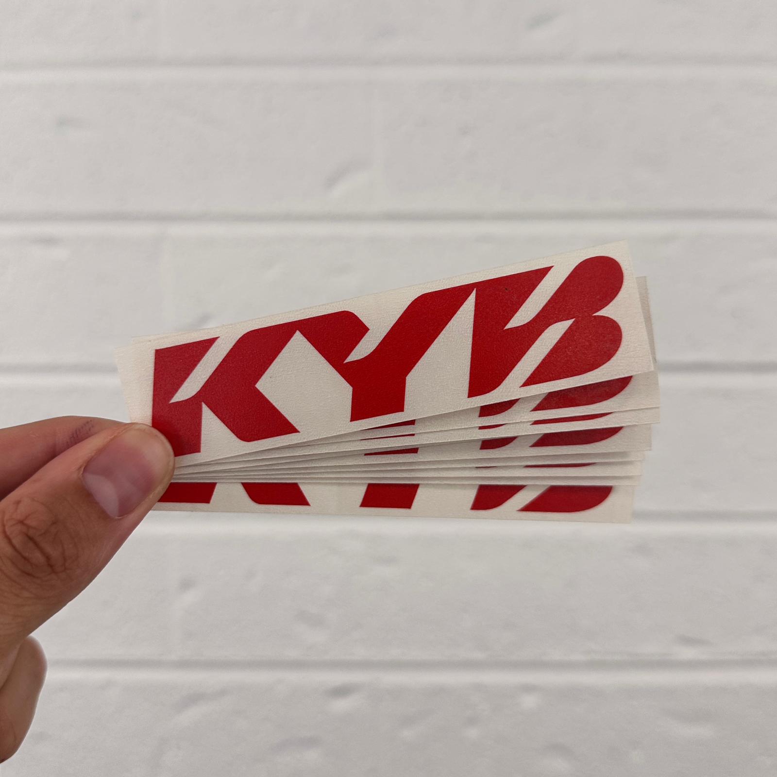 Hand holding a KYB logo sticker against a light gray background