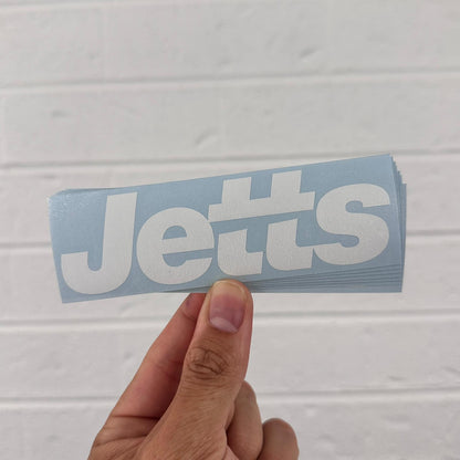 Hand holding a blue and white 'Jetts' vinyl sticker logo against a light gray background
