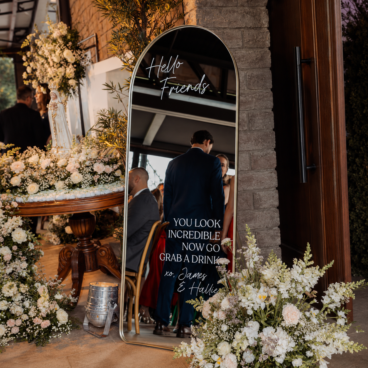 A tall arched floor mirror with a gold frame stands in a decorated event space, reflecting guests while displaying printed text that reads 'Hello Friends' and 'YOU LOOK INCREDIBLE NOW GO GRAB A DRINK' with 'xo, James & Halle' at the bottom.