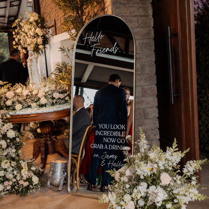 A tall arched floor mirror with a gold frame stands in a decorated event space, reflecting guests while displaying printed text that reads 'Hello Friends' and 'YOU LOOK INCREDIBLE NOW GO GRAB A DRINK' with 'xo, James & Halle' at the bottom.