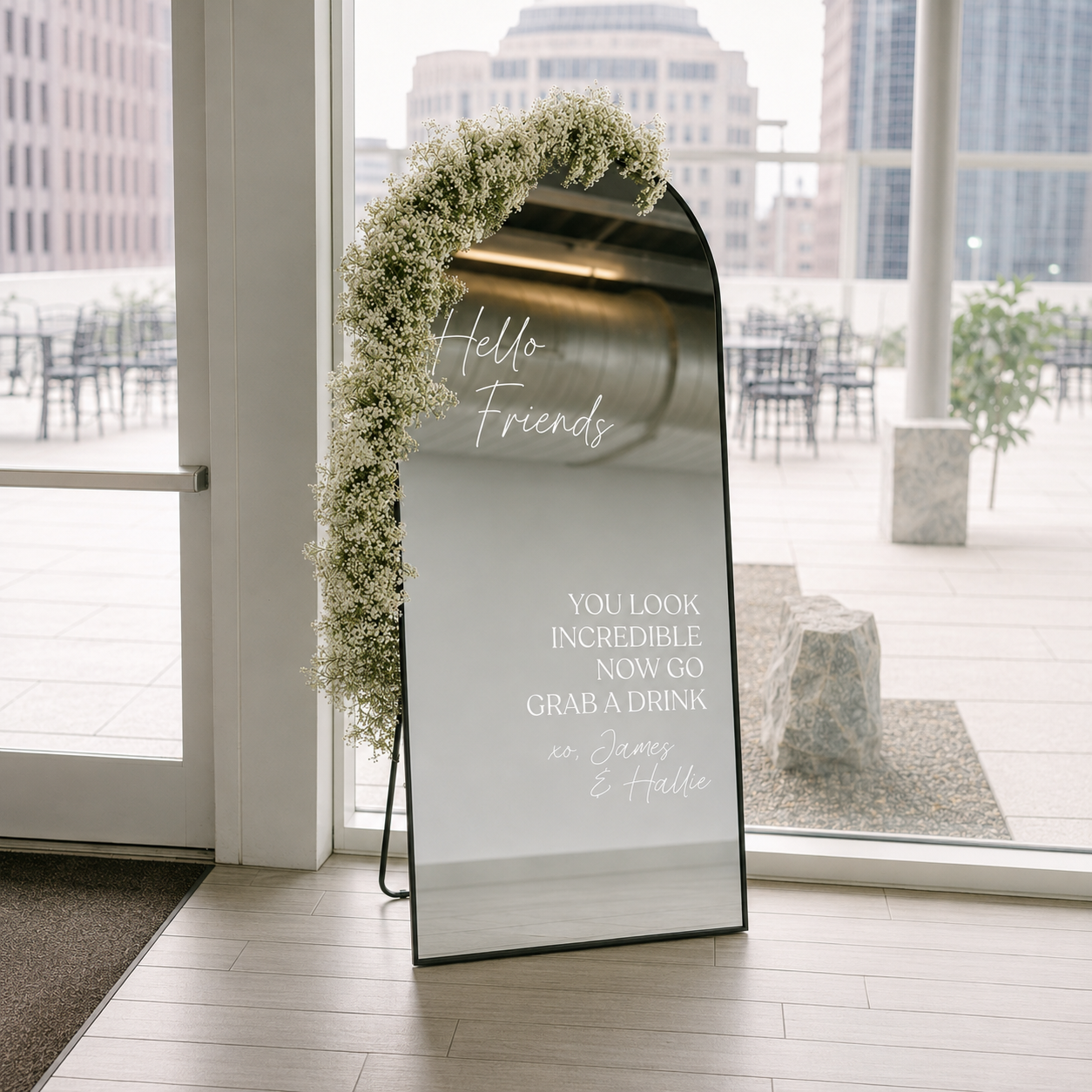 A freestanding arched mirror with a black frame and a white floral garland draped over the top, placed indoors near large windows. The mirror reflects a bright outdoor patio and is printed with wedding-style text including 'Hello Friends' and 'YOU LOOK INCREDIBLE NOW GO GRAB A DRINK'.