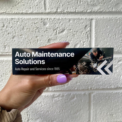 Hand holding 'Auto Maintenance Solutions' bumper sticker with 'Auto Repair and Services since 1985' text and mechanic image.