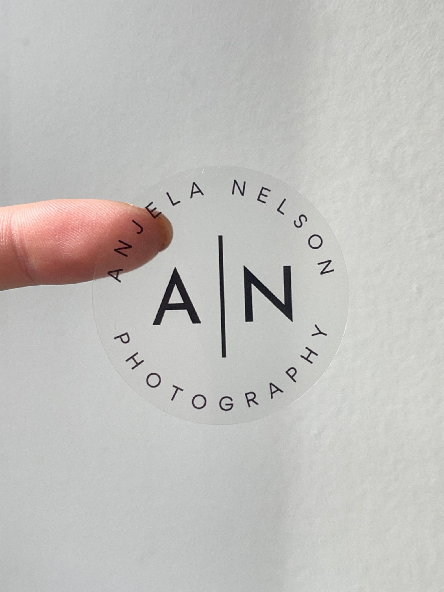 Hand holding a round transparent 'Anjela Nelson Photography' sticker with 'A | N' in the center