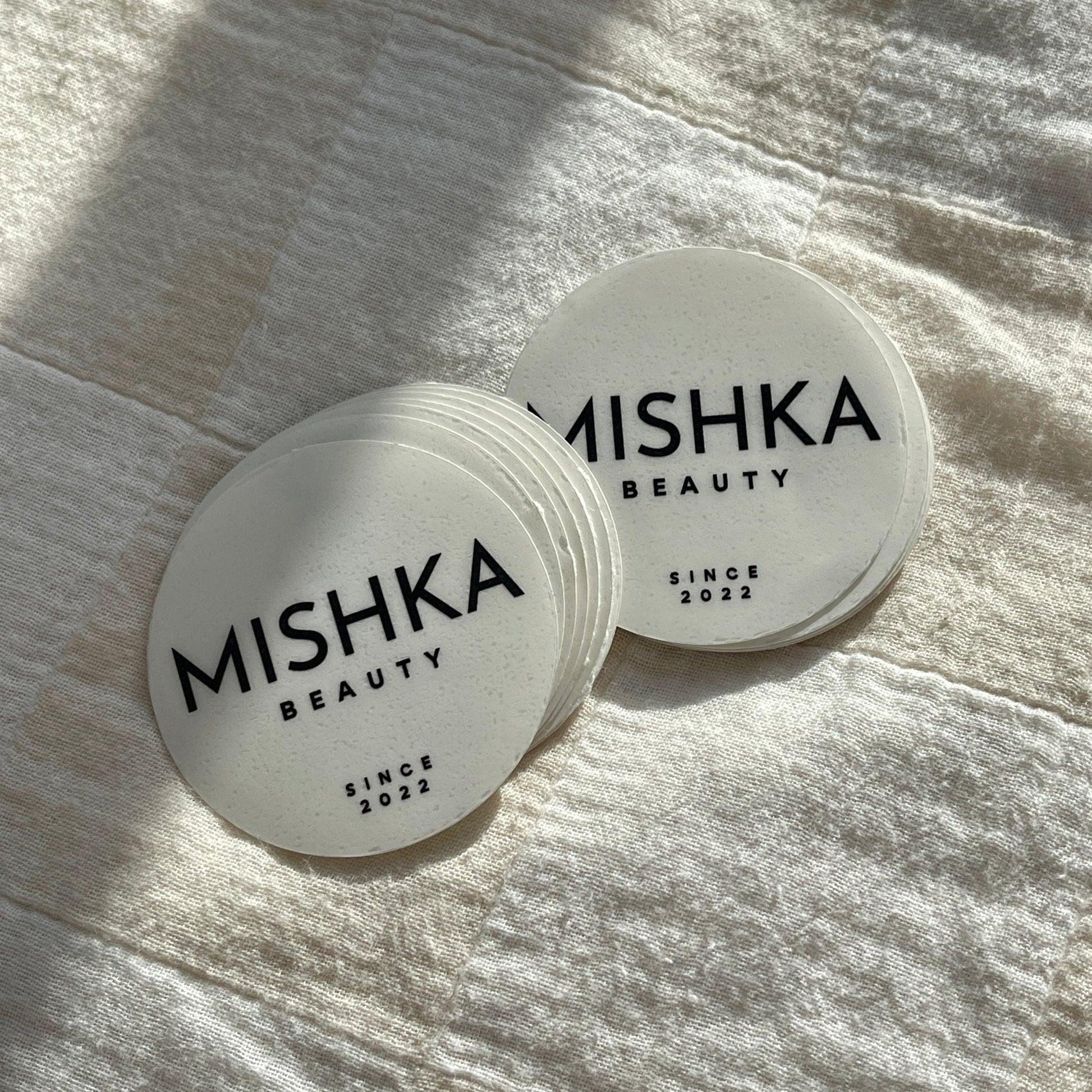 Stack of round 'Mishka Beauty' stickers with 'Since 2022' text on a textured fabric background.