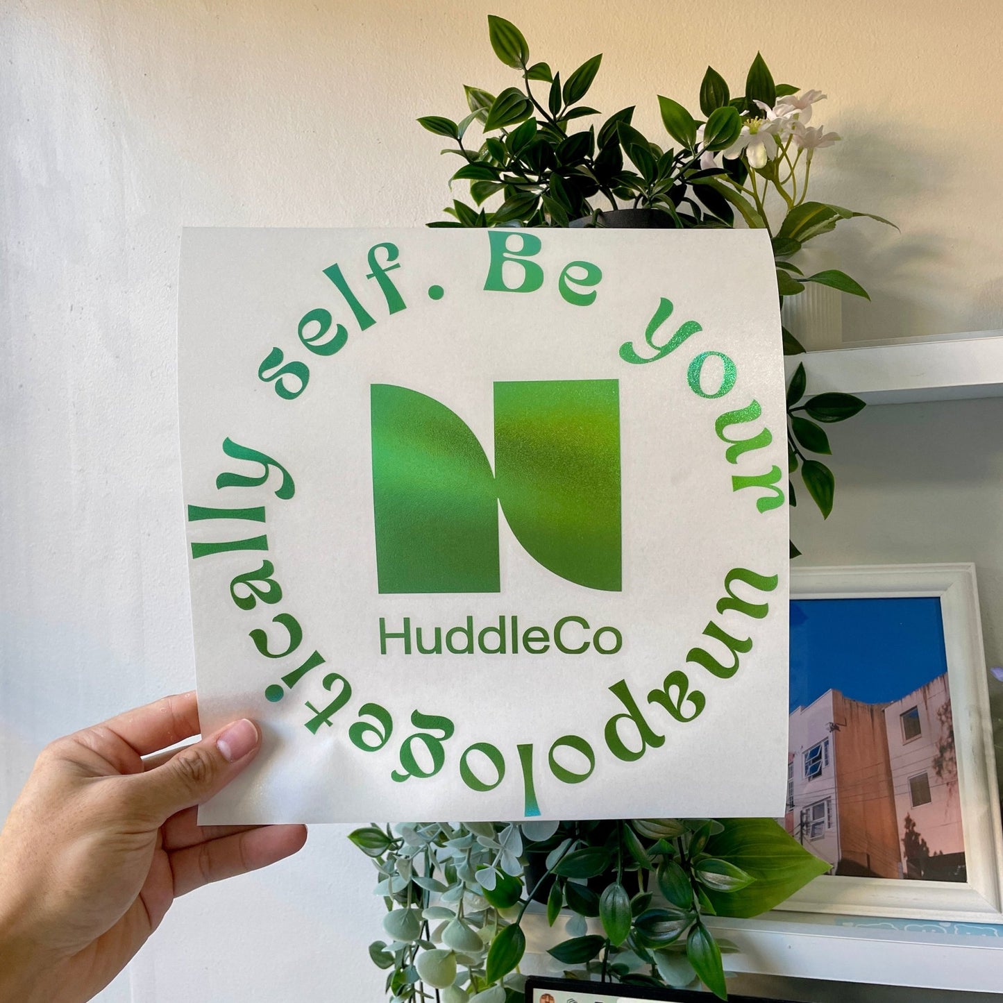 Hand holding a vinyl decal with the HuddleCo logo and the text 'Be your unapologetically self' in green, with a plant and framed picture in the background.