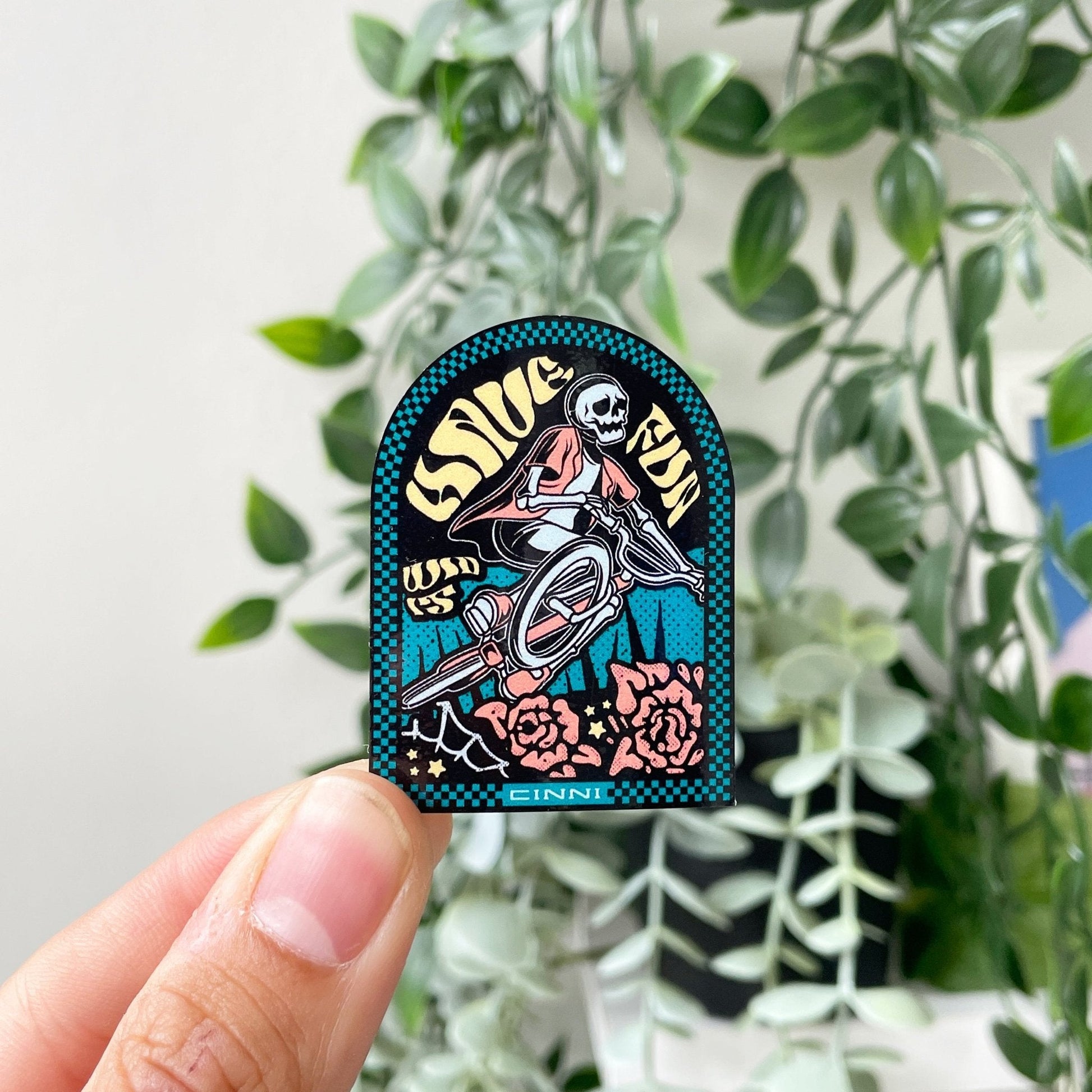 Die cut sticker featuring a skeleton riding a bicycle with the text 'Sage Run' and 'Cinni' on a colorful background, held in front of green leaves