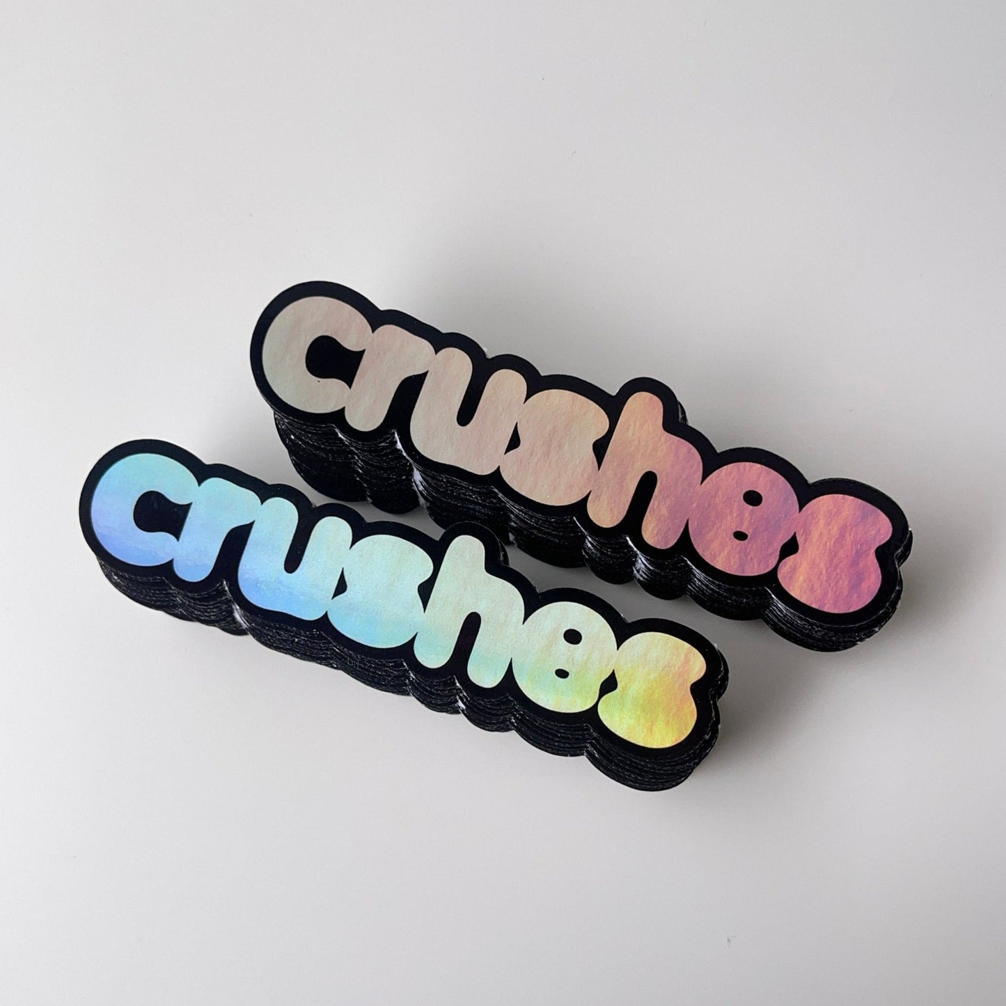 Stacks of glossy holographic die cut stickers with the text 'crushes' in a colorful gradient on a black background.