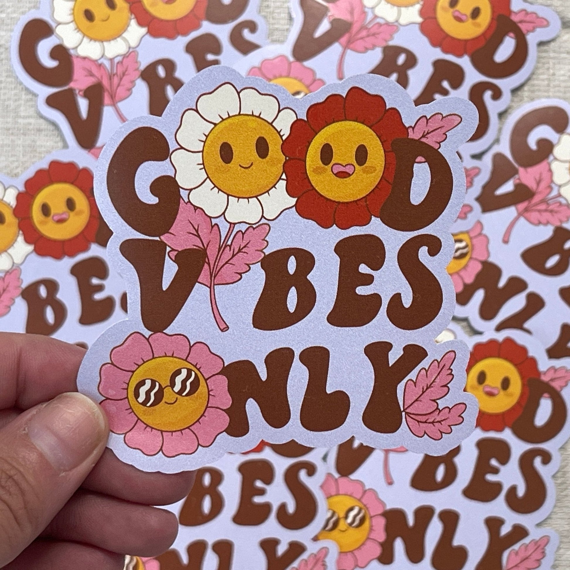 Die cut sticker with the text 'Good Vibes Only' in brown letters and illustrations of smiling flowers and pink leaves, held against a background of similar stickers.