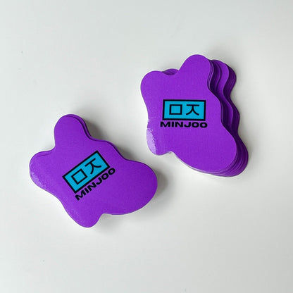 Stacks of purple gloss die cut stickers with the text 'MINJOO' in black and a blue rectangular symbol.