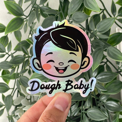 Holographic die cut sticker with a smiling cartoon child's face and the text 'Dough Baby!' held in front of green leaves.