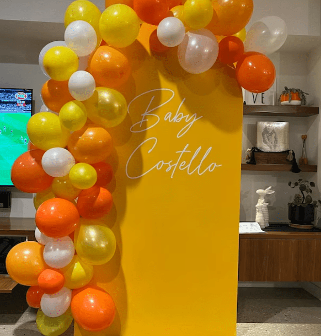 Event backdrop featuring the vinyl decal text 'Baby Costello' in white script on a yellow background, decorated with clusters of orange, yellow, and white balloons.