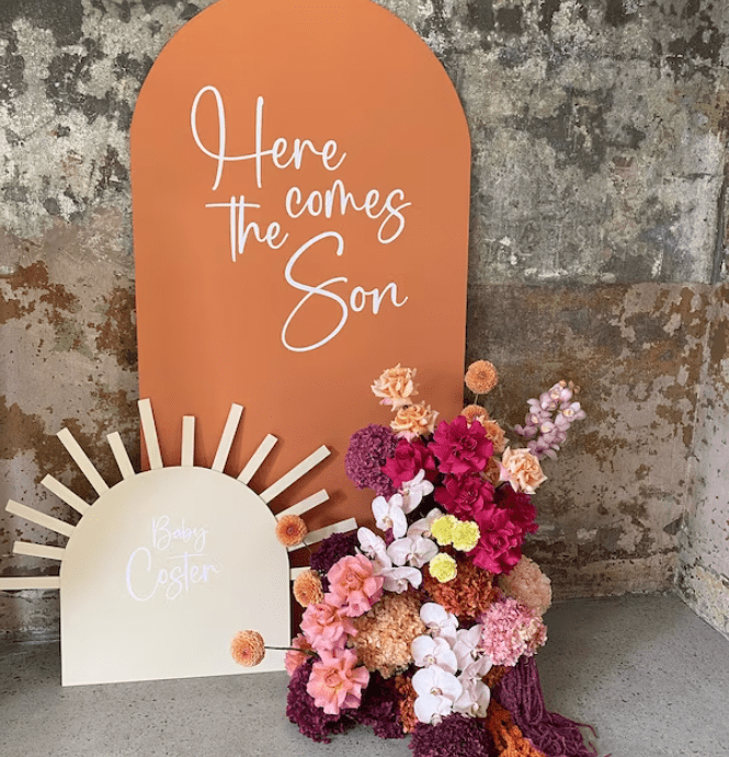 Event setup featuring a sign with 'Here comes the Son' text on an orange background, a sun-shaped decor with 'Baby Coster' text, and a colorful arrangement of flowers. The text on the signs are vinyl decals made by ntlprint.