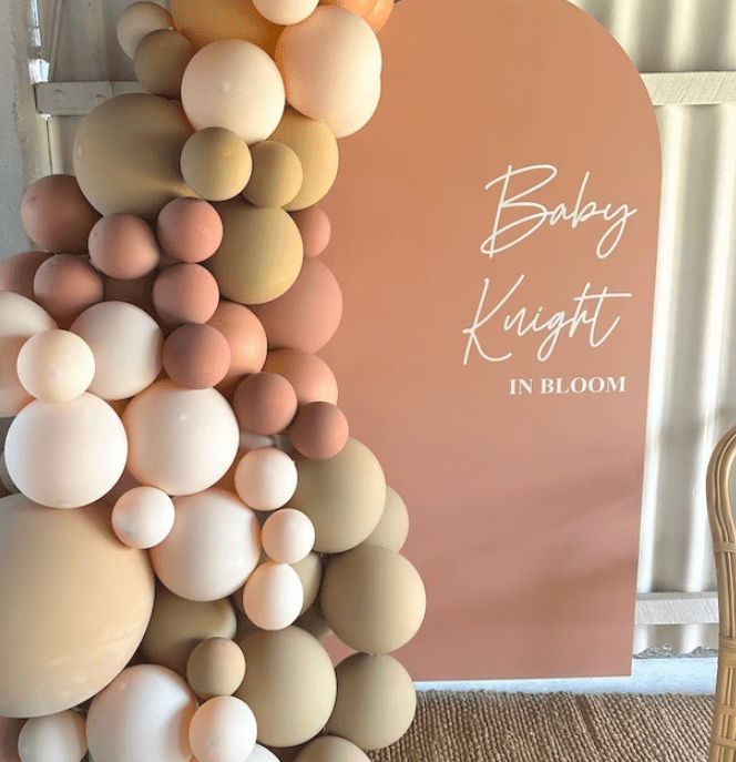 Event backdrop featuring the text 'Baby Knight In Bloom' in white script on a peach background, decorated with clusters of beige, pink, and white balloons