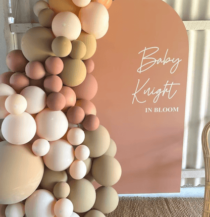 Event backdrop featuring the text 'Baby Knight In Bloom' in white script on a peach background, decorated with clusters of beige, pink, and white balloons