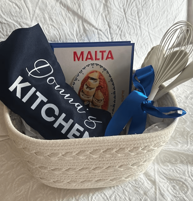 Gift basket containing a navy blue apron with 'Donna's Kitchen' text in white iron on transfer, a 'Malta' cookbook, and kitchen utensils tied with a blue ribbon