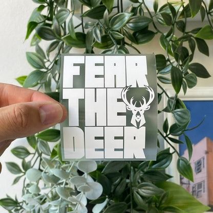 Hand holding an iron-on design with the text 'FEAR THE DEER' in bold white letters and a stylized deer head, with green leaves in the background.