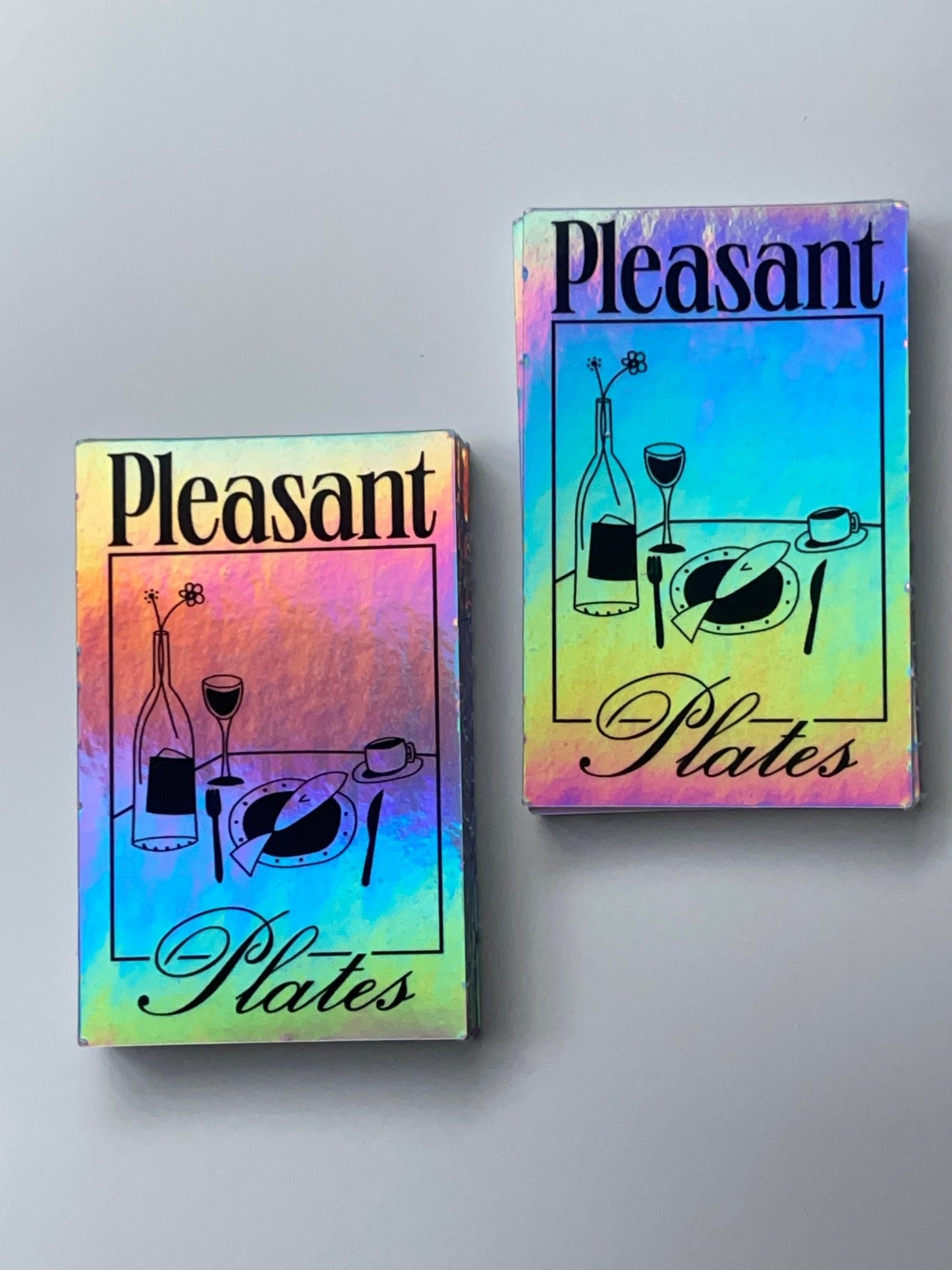 Two holographic rectangular stickers with the text 'Pleasant Plates' featuring an illustration of a dining table set with a bottle, two glasses, and plates.