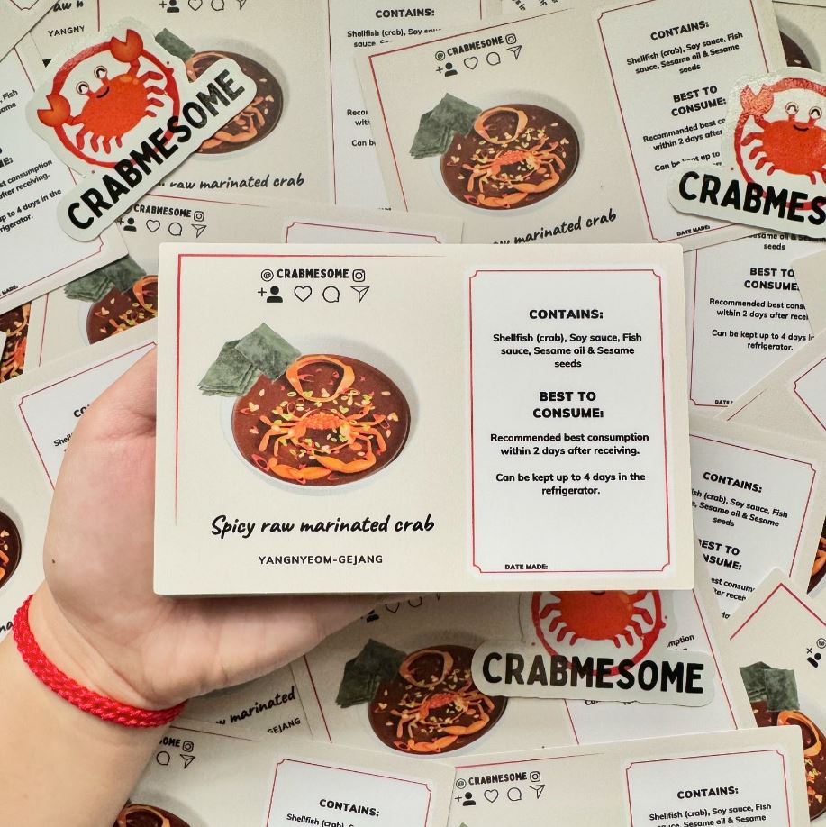 Hand holding a rectangular sticker with 'CRABMESOME' and an illustration of a spicy raw marinated crab dish, surrounded by additional stickers with similar designs and ingredient information.