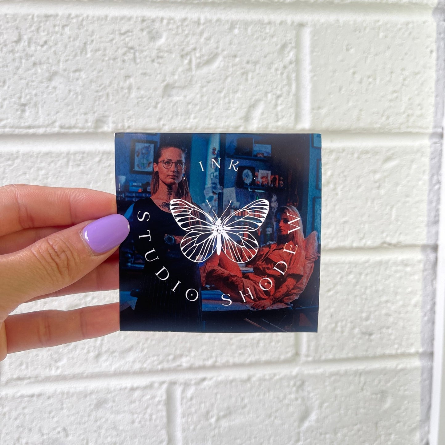 A square sticker with a butterfly logo in the center and the text "INK STUDIO SHODEV" around it. The background features an image of two individuals in a studio setting. The sticker is being held by a hand with purple nail polish.