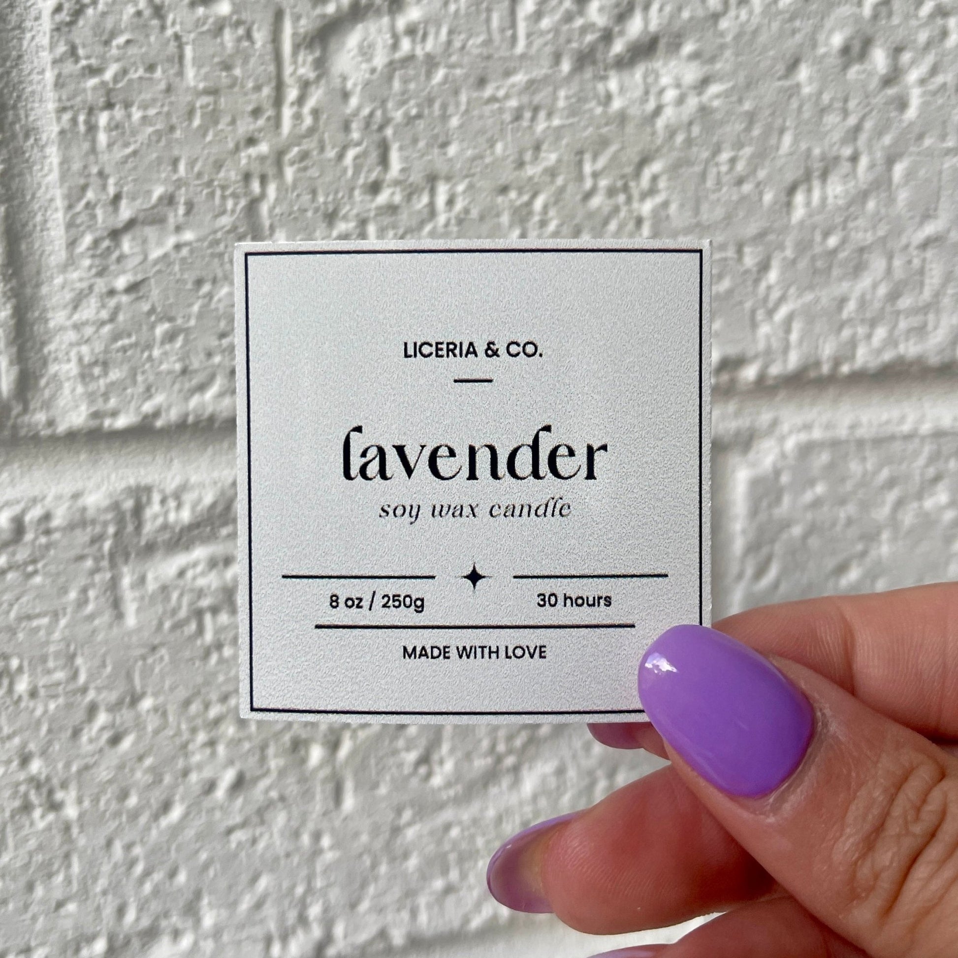 A square sticker for "Lavender soy wax candle" by Liceria & Co., with details of 8 oz / 250g and 30 hours burn time, held by a hand with purple nail polish against a white textured background.