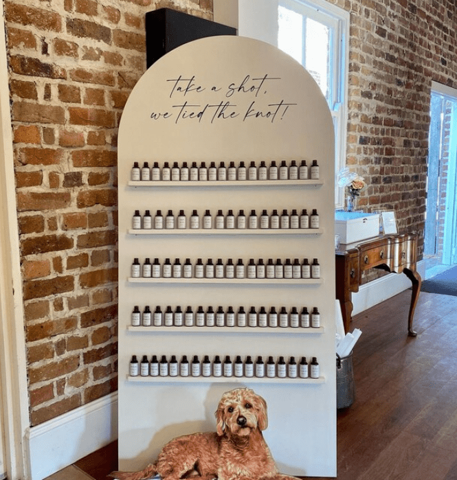 Vinyl decal on a large arched board reads "Take a shot, we tied the knot!" in cursive script, with multiple rows of small bottles beneath it and a brick wall in the background